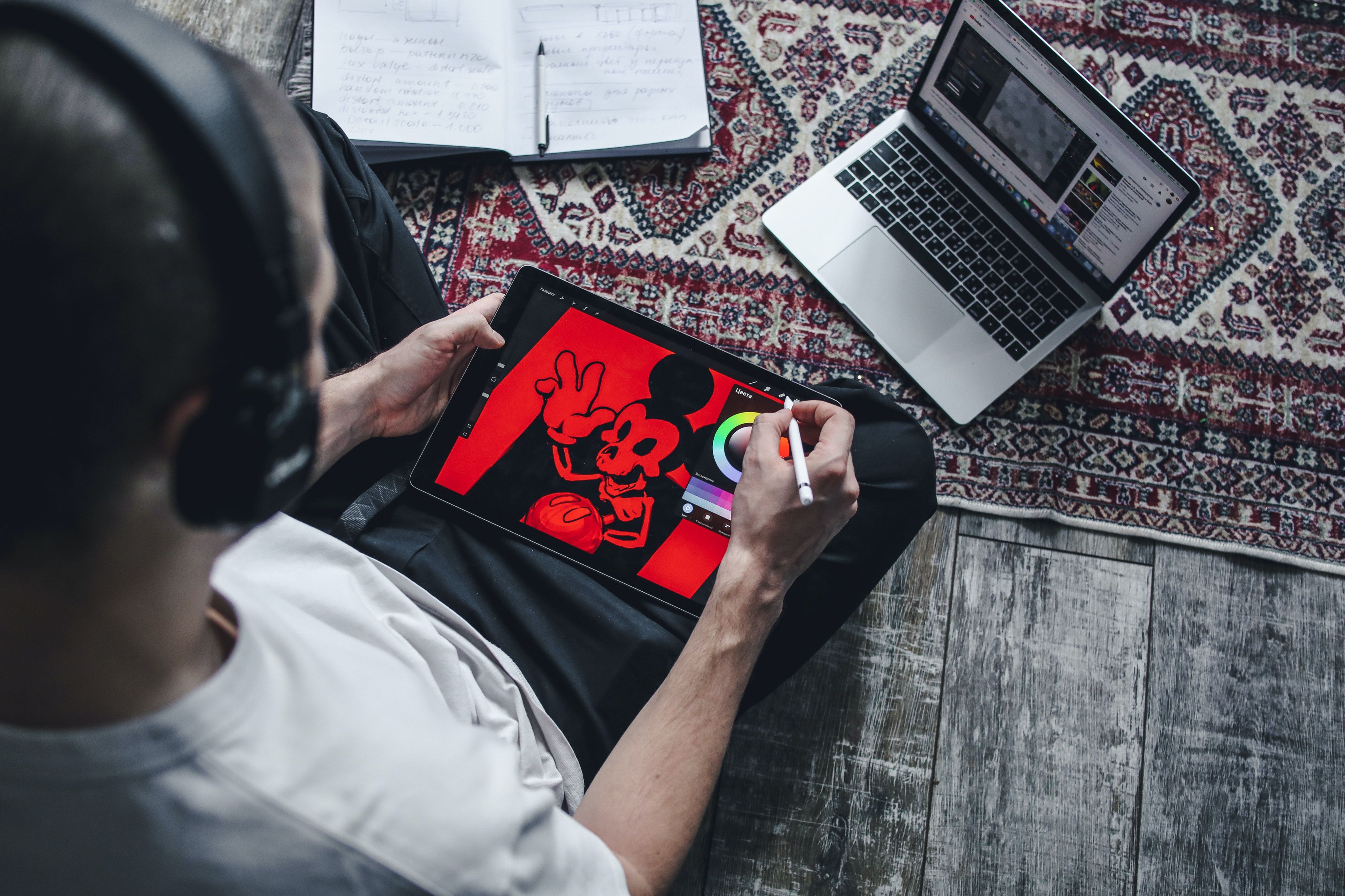 Người đàn ông ngồi trên sàn sử dụng công nghệ phác thảo phim hoạt hình trên iPad trong khi xem máy tính xách tay.