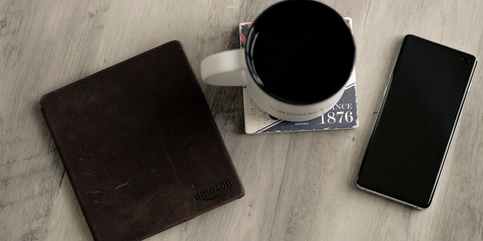10 mẹo Kindle Oasis để nhận được nhiều hơn từ thiết bị đọc sách điện tử cao cấp của bạn Photo of a Kindle oasis on a desk with a smartphone and a mug